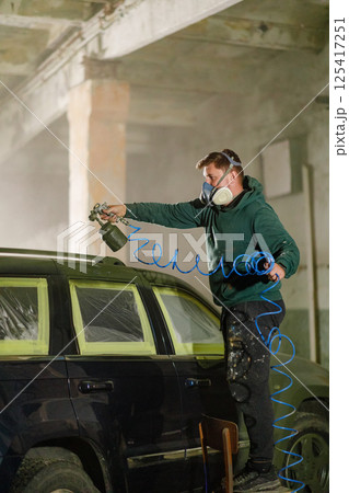 A man in a green hoodie wears a mask and goggles while using a spray gun to paint a vehicle inside a workshop. Protective tape covers parts of the car for precision work. A man in a green hoodie wears a mask and goggles while using a spray gun to paint a vehicle inside a workshop. Protective tape covers parts of the car for precision work. 125417251