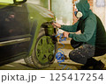 A person wearing a hooded sweatshirt and mask is spray painting a car wheel in a dimly lit garage. Tools and hoses surround the area, emphasizing the DIY project atmosphere. 125417254
