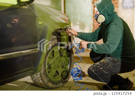 A person wearing a hooded sweatshirt and mask is spray painting a car wheel in a dimly lit garage. Tools and hoses surround the area, emphasizing the DIY project atmosphere. A person wearing a hooded sweatshirt and mask is spray painting a car wheel in a dimly lit garage. Tools and hoses surround the area, emphasizing the DIY project atmosphere. 125417254