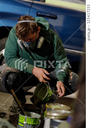 A young man, dressed in a green hoodie and protective gear, carefully pours paint onto the ground while crouched beside a parked vehicle. 125417255
