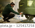 A technician wearing a mask sprays green paint on the rear of a vehicle in a cluttered workshop. The area is filled with tools and equipment, emphasizing the restoration work in progress. 125417258