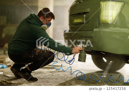 A technician wearing a mask sprays green paint on the rear of a vehicle in a cluttered workshop. The area is filled with tools and equipment, emphasizing the restoration work in progress. A technician wearing a mask sprays green paint on the rear of a vehicle in a cluttered workshop. The area is filled with tools and equipment, emphasizing the restoration work in progress. 125417258