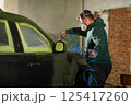 A worker focuses on restoring a car in a workshop, applying paint with precision while wearing safety gear. The environment is filled with tools and projects underway, highlighting craftsmanship. 125417260