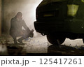 In a dimly lit urban area at night, an individual wearing a gas mask is crouching next to a vehicle, seemingly focused on a task involving tools and equipment. The atmosphere is tense and mysterious. 125417261