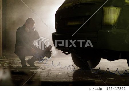 In a dimly lit urban area at night, an individual wearing a gas mask is crouching next to a vehicle, seemingly focused on a task involving tools and equipment. The atmosphere is tense and mysterious. In a dimly lit urban area at night, an individual wearing a gas mask is crouching next to a vehicle, seemingly focused on a task involving tools and equipment. The atmosphere is tense and mysterious. 125417261