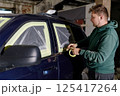 A man in a green hoodie meticulously applies masking tape around the windows of a dark blue SUV in a workshop. The process occurs in an industrial setting under dim lighting. 125417264