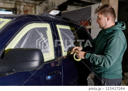 A man in a green hoodie meticulously applies masking tape around the windows of a dark blue SUV in a workshop. The process occurs in an industrial setting under dim lighting. 125417264