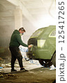 A man wears a mask while spray painting a vehicle inside a dimly lit workshop filled with dust and debris. He focuses on the car's rear, engaged in a detailed restoration project. 125417265