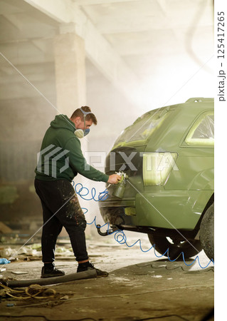 A man wears a mask while spray painting a vehicle inside a dimly lit workshop filled with dust and debris. He focuses on the car's rear, engaged in a detailed restoration project. A man wears a mask while spray painting a vehicle inside a dimly lit workshop filled with dust and debris. He focuses on the car's rear, engaged in a detailed restoration project. 125417265