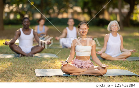 Multinational yoga enthusiasts with female coach perform padmasana in park 125418894
