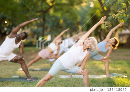Elderly woman practices yoga pose Anjaneyasana with other people of different nationalities in summer park 125418925