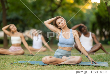 Multinational yoga enthusiasts with female coach perform padmasana in park Multinational yoga enthusiasts with female coach perform padmasana in park 125418996