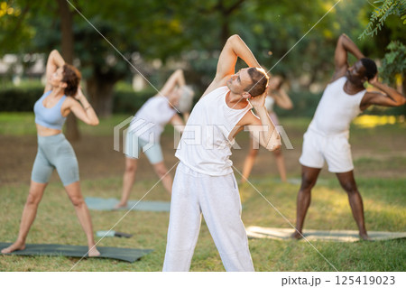 Man performing Bending Tree Pose during group yoga in park Man performing Bending Tree Pose during group yoga in park 125419023