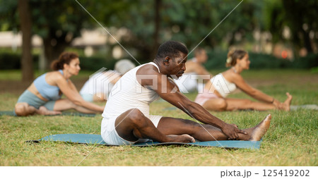 Multinational yoga enthusiasts with African male coach perform Janu Shirshasana in park 125419202