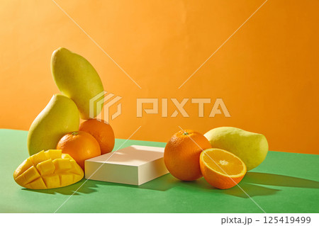 Mangoes are stacked vertically on top of round oranges, surrounded by mango slices with evenly spaced squares. A rectangular white pedestal is placed between the fruits. Empty space for text.  125419499