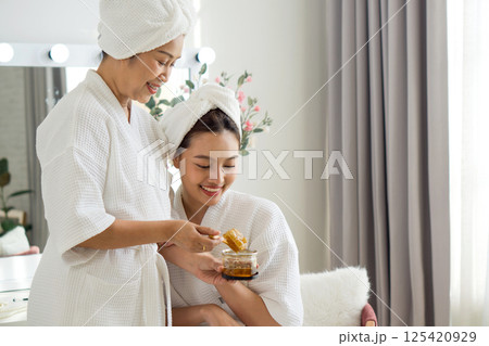 Asian mother and daughter share a joyful moment as they apply skincare product in a cozy home spa. They are dressed in comfortable robe and towel, surrounded by a tranquil atmosphere. 125420929