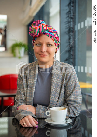 Young woman in a turban sitting in a cafe, drinking coffee and enjoying a peaceful moment. The concept shows that people with illness continue to live, socialize, and express their individuality 125420960