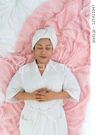 Senior asian woman dressed in a cozy white robe lies peacefully on a soft pink blanket with her hand resting on her stomach. The atmosphere promotes tranquility and self-care. 125421047