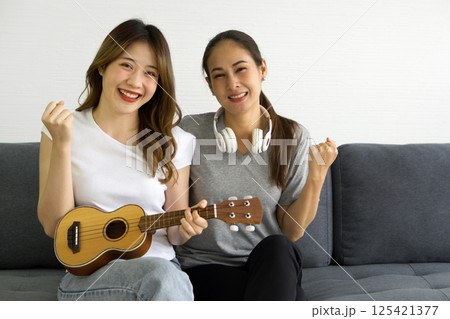 Two asian women playing Ukulele guitar, sitting on sofa in the living room. Enjoy leisure weekend at home. Stress free concept. 125421377