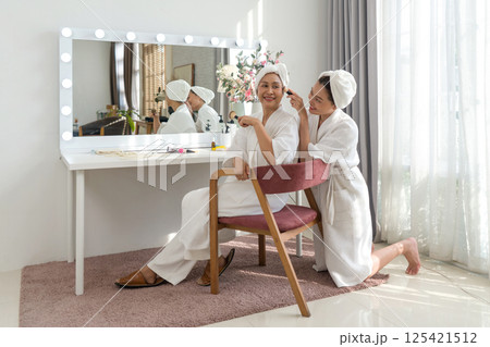 Asian mother and daughter share a joyful moment as they apply skincare product with a brush in cozy home spa. They are dressed in comfortable robe and towel, sit in front of a vanity mirror with light 125421512
