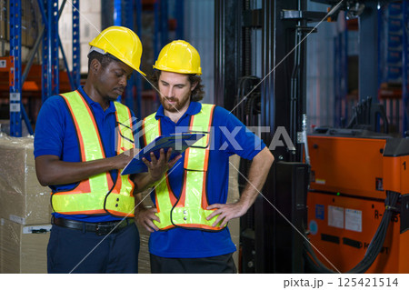 Two warehouse worker in yellow hard hat and safety vest discuss operational detail while examining document in an industrial setting. They focus intently on their task. Two warehouse worker in yellow hard hat and safety vest discuss operational detail while examining document in an industrial setting. They focus intently on their task. 125421514