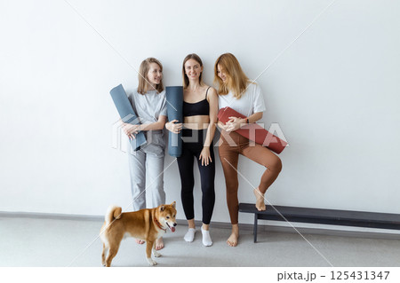 Beautiful young girls in their hands with a yoga mat or karemat stand against the wall smiling before training, and with them a dog indoors Beautiful young girls in their hands with a yoga mat or karemat stand against the wall smiling before training, and with them a dog indoors 125431347