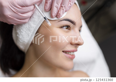Close up of hands of young cosmetologist injecting botox in female face. She is standing and smiling. The woman is closed her eyes with relaxation Close up of hands of young cosmetologist injecting botox in female face. She is standing and smiling. The woman is closed her eyes with relaxation 125431360