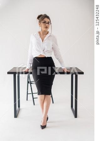 Confident businesswoman posing with hands on desk in office setting wearing white shirt and black skirt 125432248