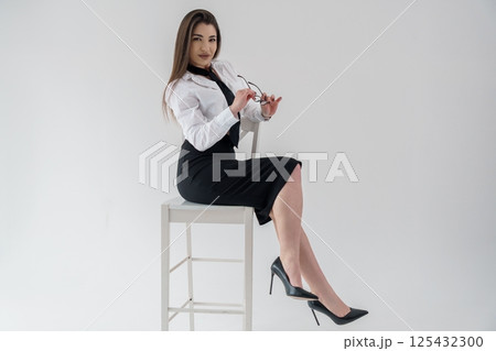 Attractive businesswoman sitting on white chair, adjusting glasses, wearing black dress and white shirt 125432300