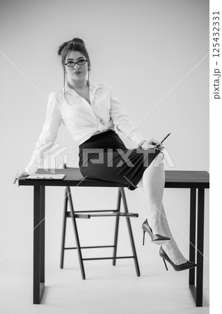 Elegant businesswoman sitting on desk, confidently posing in a black and white portrait Elegant businesswoman sitting on desk, confidently posing in a black and white portrait 125432331