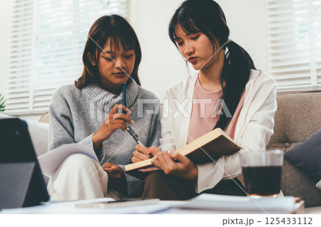 Female higher education student using laptop and studying book in home, Online learning. 125433112