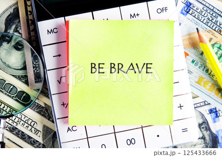 A bright yellow sticky note with the message Be Brave rests on a calculator. Surrounding it are various denominations of money and office supplies, creating an inspiring workspace 125433666