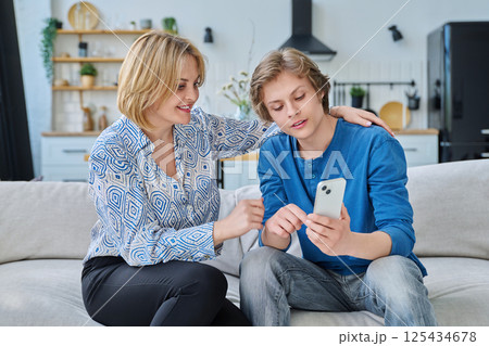 Middle aged mother and teenage son sitting together at home looking at smartphone Middle aged mother and teenage son sitting together at home looking at smartphone 125434678