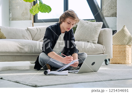 Teenager guy student studying sitting at home on the floor using laptop textbook notebook 125434691