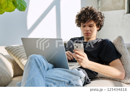Handsome young man with laptop using smartphone sitting on sofa at home 125434818
