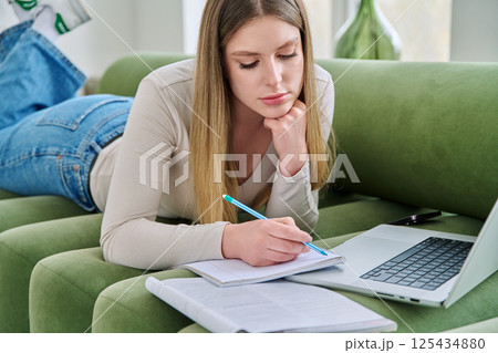 Young student girl lying at home on couch with laptop, notebook, textbook 125434880