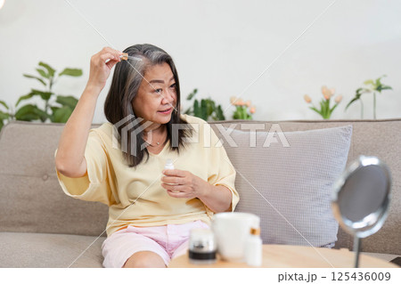 Beauty and Age Diversity. A senior woman applying hair serum with confidence in her living room. 125436009
