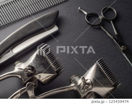 Various grooming tools including clippers, scissors, and a comb are arranged on a dark surface, showcasing the essentials for hair cutting in a barbershop environment. Various grooming tools including clippers, scissors, and a comb are arranged on a dark surface, showcasing the essentials for hair cutting in a barbershop environment. 125439474