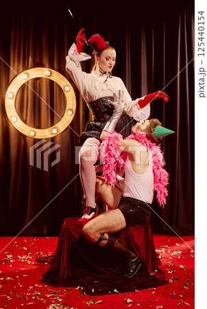 Playful cabaret scene with dominant woman actress in top hat and male performer kneeling on red velvet. 125440154