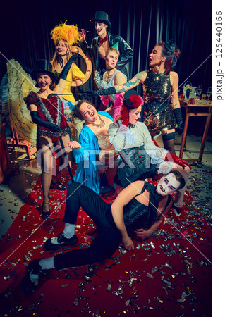 Cabaret troupe laughing and posing in exaggerated gestures under stage lights with expressive costumes and red carpet. 125440166