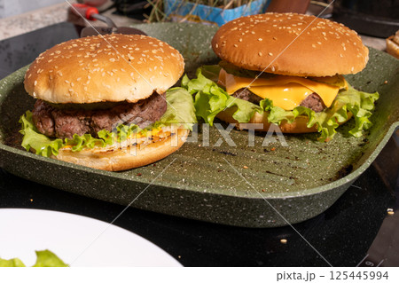 Two burgers with sesame buns, beef patties, lettuce, melted cheese, and sauce on a green grill pan, perfect for homemade meal scenes. Two burgers with sesame buns, beef patties, lettuce, melted cheese, and sauce on a green grill pan, perfect for homemade meal scenes. 125445994