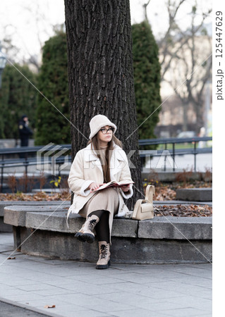 Sitting against a tree in a park, a young woman enjoys a book while dressed in cozy winter attire during a quiet afternoon in an urban setting 125447629