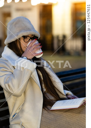 Cozy evening with a warm drink and a good book while sitting on a park bench under soft glowing lights in a charming urban setting Cozy evening with a warm drink and a good book while sitting on a park bench under soft glowing lights in a charming urban setting 125447631