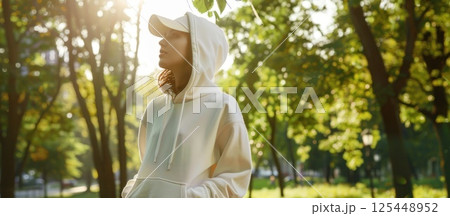 Young woman in a white hoodie and cap standing in a park on a sunny day. 125448952