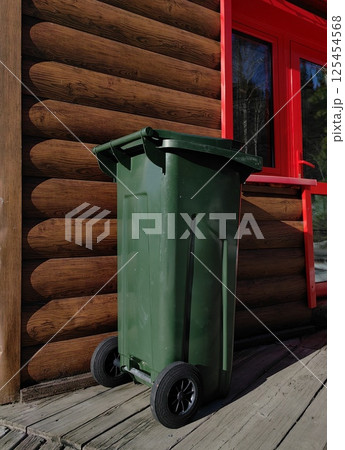 green trash bin for recycling positioned near wooden cabin with bright red window. green trash bin for recycling positioned near wooden cabin with bright red window. 125454568