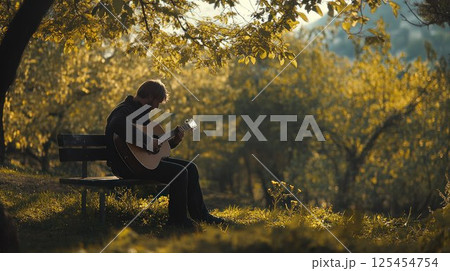 Man plays acoustic guitar on park bench under autumn trees. Man plays acoustic guitar on park bench under autumn trees. 125454754