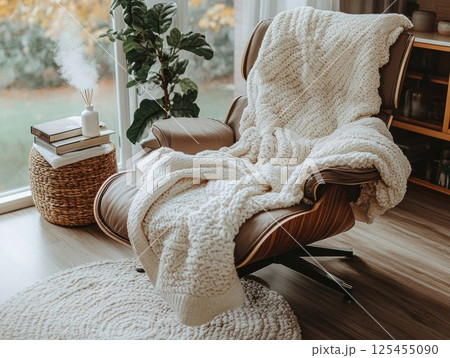 Cozy cream knit blanket draped over modern armchair, autumnal window view, home interior 125455090
