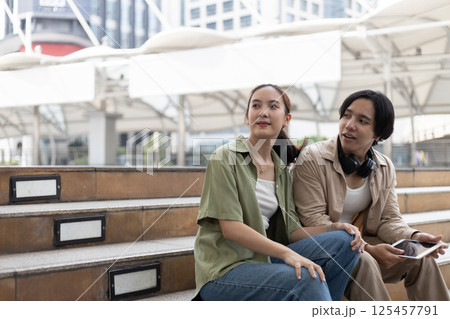 Two young adult engage in a relaxed conversation on stepped seating in a contemporary urban environment, surrounded by buildings and natural light during the day. 125457791