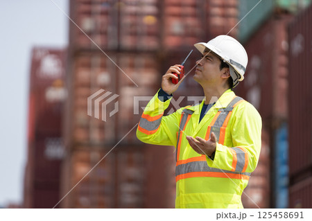 container loading worker in fluorescent safety jacket and hard hat is using walkie-talkie radio to communicate while surrounded stacked shipping containers. His role involves coordinating logistics. 125458691