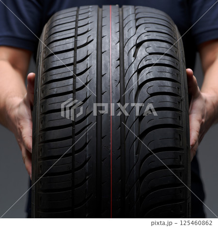 A pair of hands carefully hold a modern car tire, showing off the tread pattern. The focus is sharply on the center of the tread, with the hands slightly out of focus in the foreground and bac 125460862